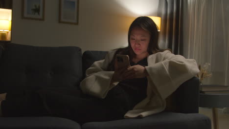 woman spending evening at home sitting on sofa with mobile phone scrolling through internet or social media 8