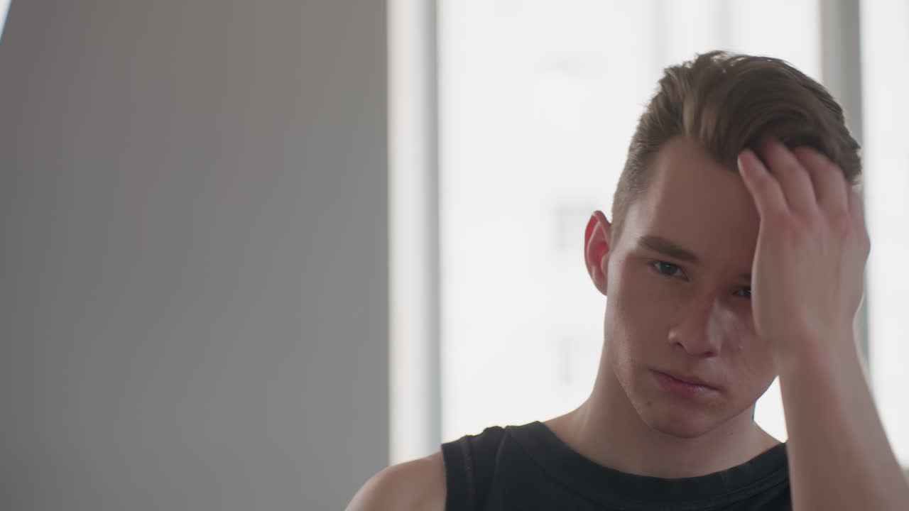 handsome young student in black tank top stands before bright window mirror, confidently runs fingers through styled hair, showcasing focused self care moment and modern grooming routine