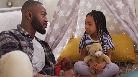 happy african american father and daughter sitting in tent and telling fairy tales