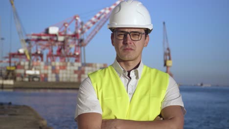 retrato de un joven inspector de control serio con gafas y un casco blanco de pie frente al puerto de carga de contenedores