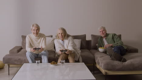 two happy senior women playing video game sitting on sofa at home while enthusiastic elderly man lying on sofa watching them and eating something