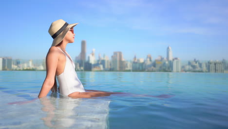 sexy asian woman in swimsuit sitting on rooftop infinity pool edge with stunning view of modern metropolis in background