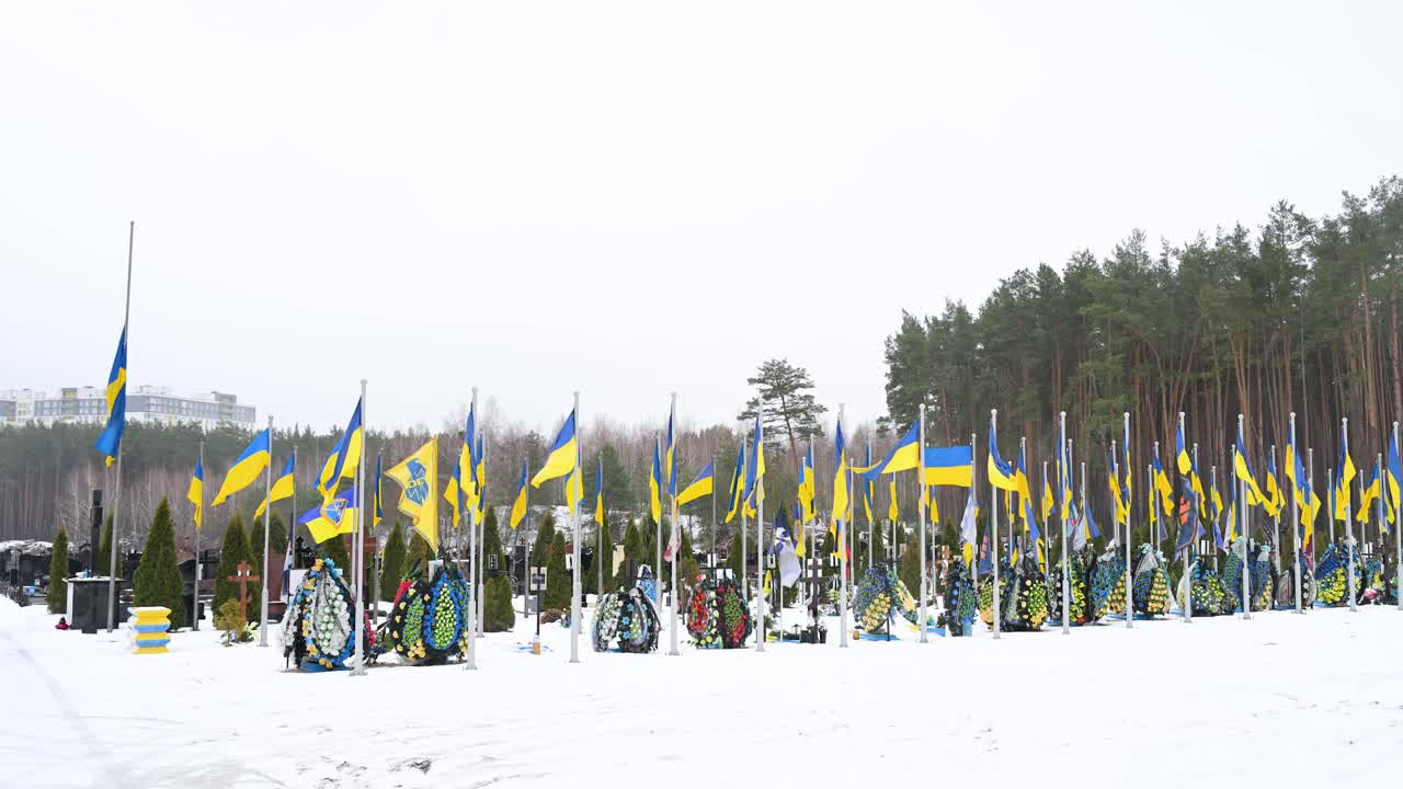 Wide shot of Irpin cemetery on a winter day, where Ukrainian soldiers who fell in the Ukraine-Russia war are laid to rest, with national flags fluttering in the wind.