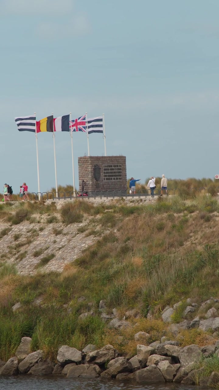 Pedestrians walk along coastal pathway by memorial monument, flags waving, under natural daylight, wide shot