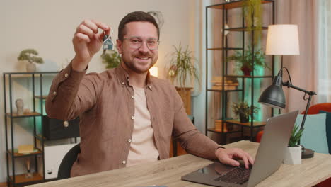 happy businessman real estate agent using laptop at home office desk showing the keys of new house