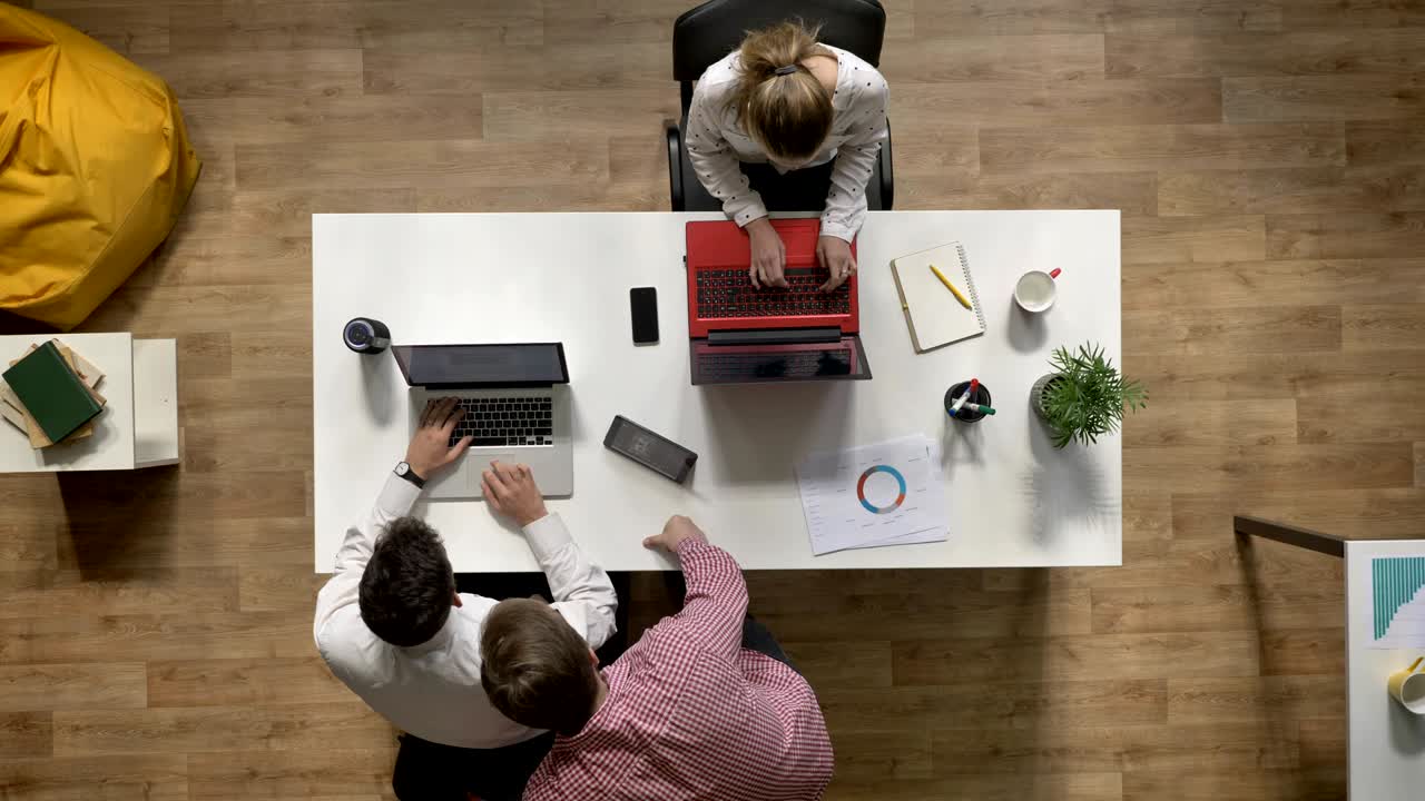 To forretningsmænd taler over bærbar computer, mens en kvinde arbejder og sidder ved et bord på et moderne kontor, topshot, arbejdskoncept