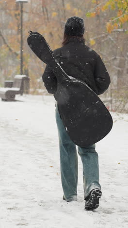 vista trasera de alguien con gorra negra y sudadera con capucha con las manos en el bolsillo, llevando una guitarra en el paquete, caminando en la nieve, banco y árboles en el fondo con una paloma en la distancia
