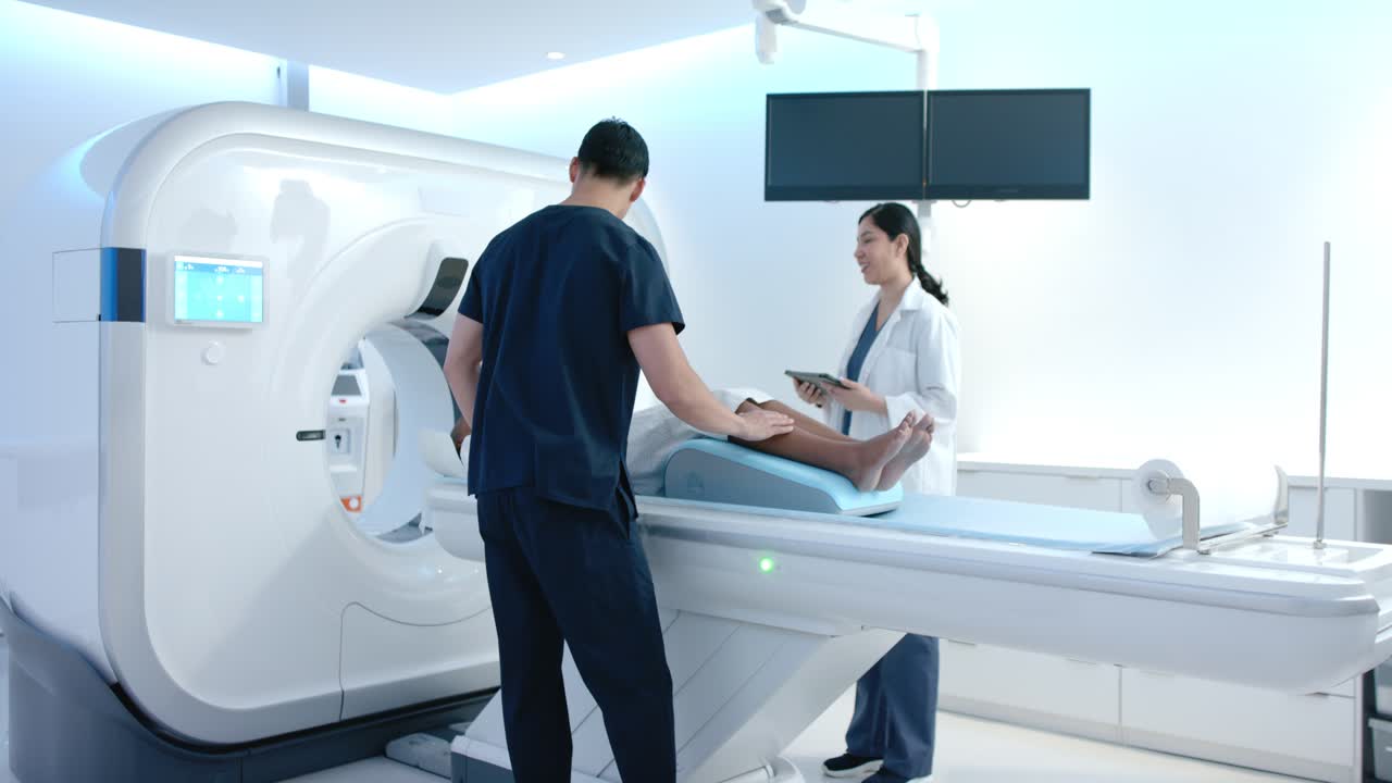 Personale sanitario vario in camice chirurgico posiziona un paziente afroamericano su un materassino dopo un controllo con tablet, muovendosi verso lo scanner.