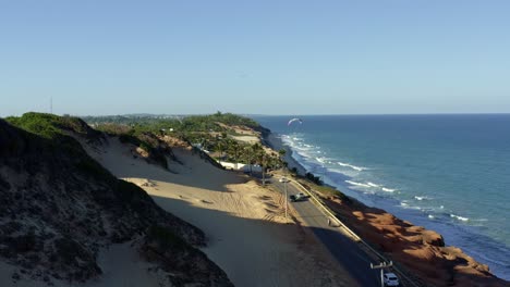 Aerial-drone-shot-of-a-paraglider-flying-down-the-famous-tropical-Cacimbinhas-Cliffs-near-Pipa,-Brazil-in-Rio-Grande-do-Norte-with-large-sand-dunes,-green-foliage,-and-crystal-blue-ocean-water