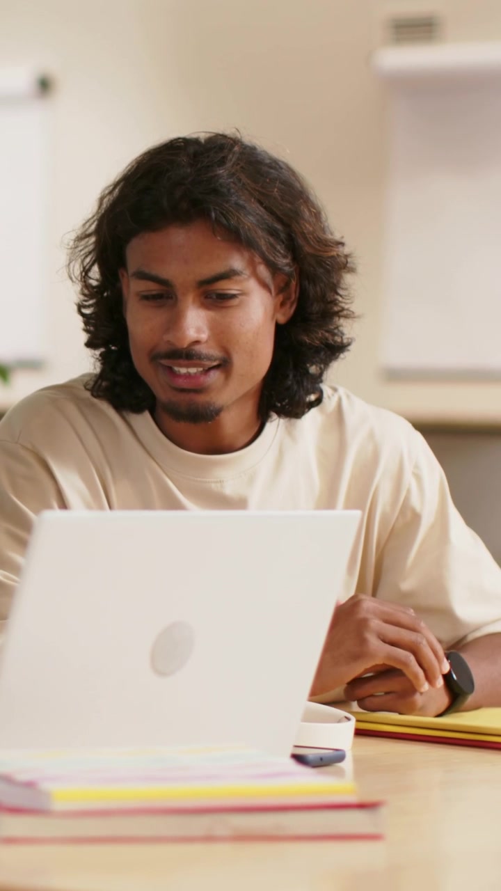 Vertical video: Beige-tee man responding to offscreen voice at study table, using laptop, notebooks