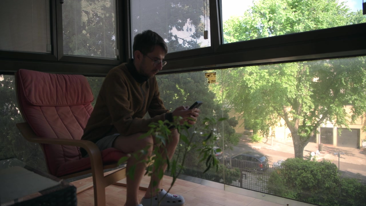 Un uomo è seduto su un balcone interno, concentrato sul suo smartphone. Attraverso grandi finestre, è visibile una vista di alberi frondosi e una strada sottostante. Adatto per contenuti tech o lifestyle.