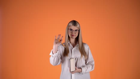 una mujer rubia bonita sosteniendo un libro y un lápiz, agitando la mano un gesto de saludo, una toma de estudio colorida