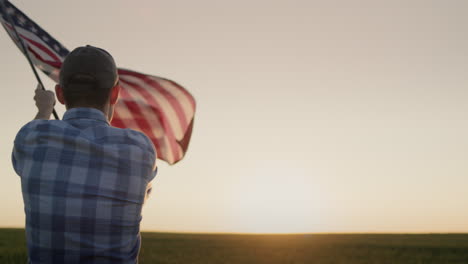 silhouette of a young man waving the american flag. standing in a field of wheat at sunset. back view