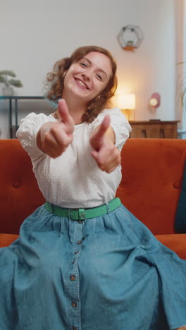 portrait of excited smiling caucasian woman showing double thumbs up sitting on sofa couch at home