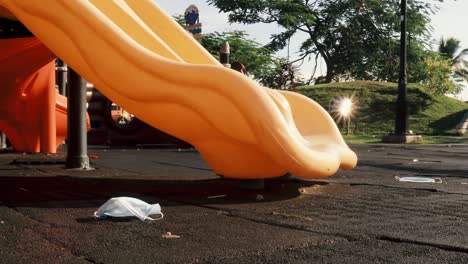 a dolly shot of a plastic orange children slide in a deserted public playground, surgical facemasks lay littered around the park