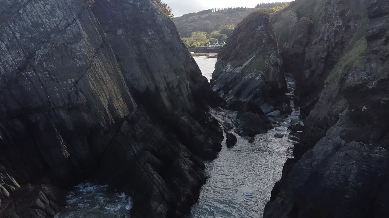 Aerial drone flying through rock formations in the sea to reveal a coastal town in a valley on the shoreline - Lee Bay, Beach, Ilfracombe, Devon, England
