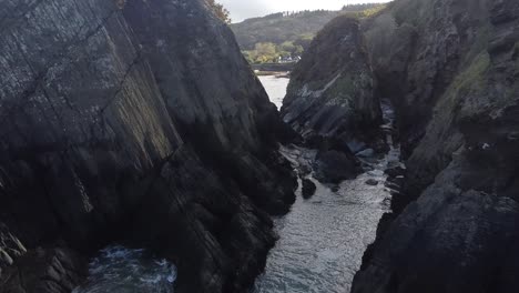 Aerial-drone-flying-through-rock-formations-in-the-sea-to-reveal-a-coastal-town-in-a-valley-on-the-shoreline---Lee-Bay,-Beach,-Ilfracombe,-Devon,-England