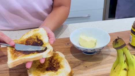 person spreads butter and dark yeast extract onto slices of white bread toast on a wooden board in a brightly lit kitchen