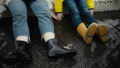 Close-up-shot-of-a-woman-in-black-rubber-boots-and-her-teenage-daughter-in-orange-rubber-boots-putting-their-feet-on-a-puddle-after-rain-on-the-street