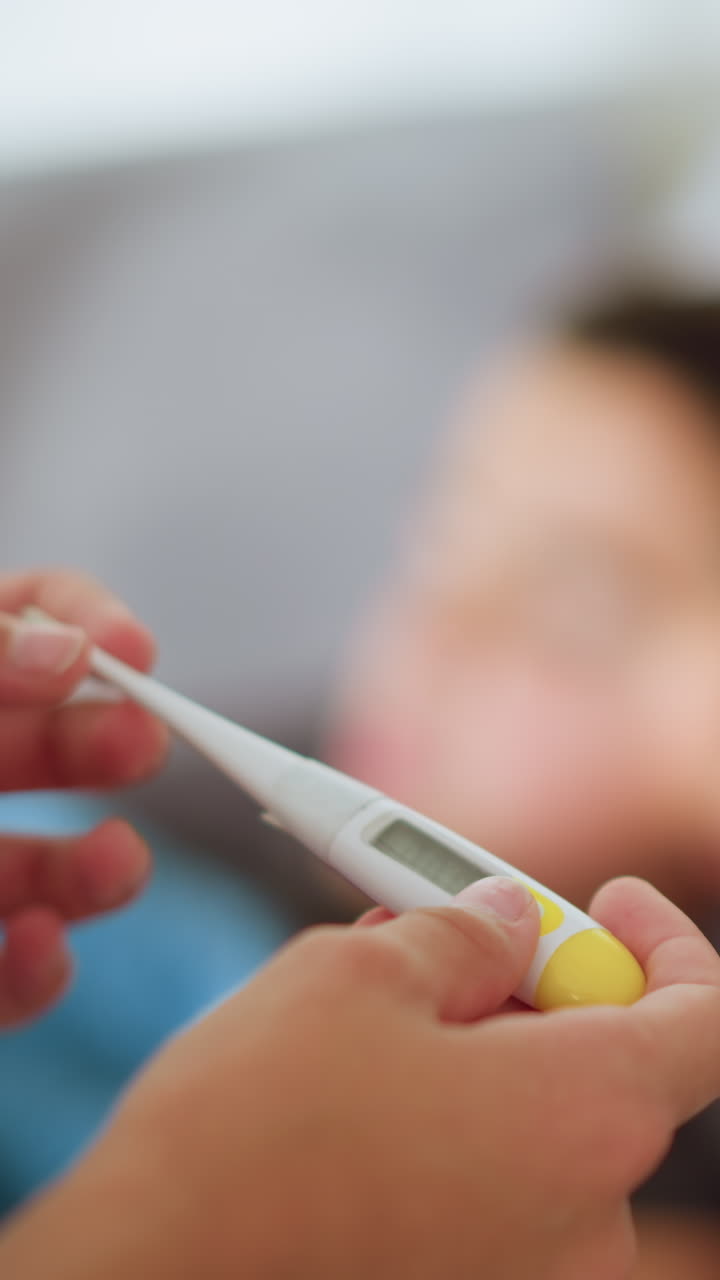 Primo piano di una persona che controlla un termometro con una visuale sfocata di una persona sdraiata sullo sfondo, ambiente interno, preoccupazione per la salute, controllo della temperatura, cura dei bambini, monitoraggio della salute