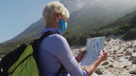 Ältere-Wanderfrau-Mit-Gesichtsmaske-Und-Rucksack,-Die-Beim-Wandern-Am-Strand-Karten-Liest.