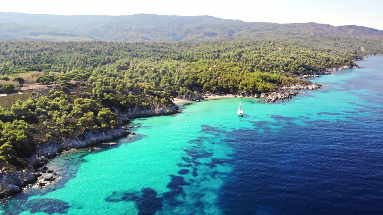 Vista aerea da drone della costa rocciosa del Mar Egeo con acqua blu trasparente, vegetazione varia, colline e barca a vela ormeggiata. Grecia
