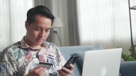 asian man sitting on couch in living room holding credit card and using smartphone shopping online