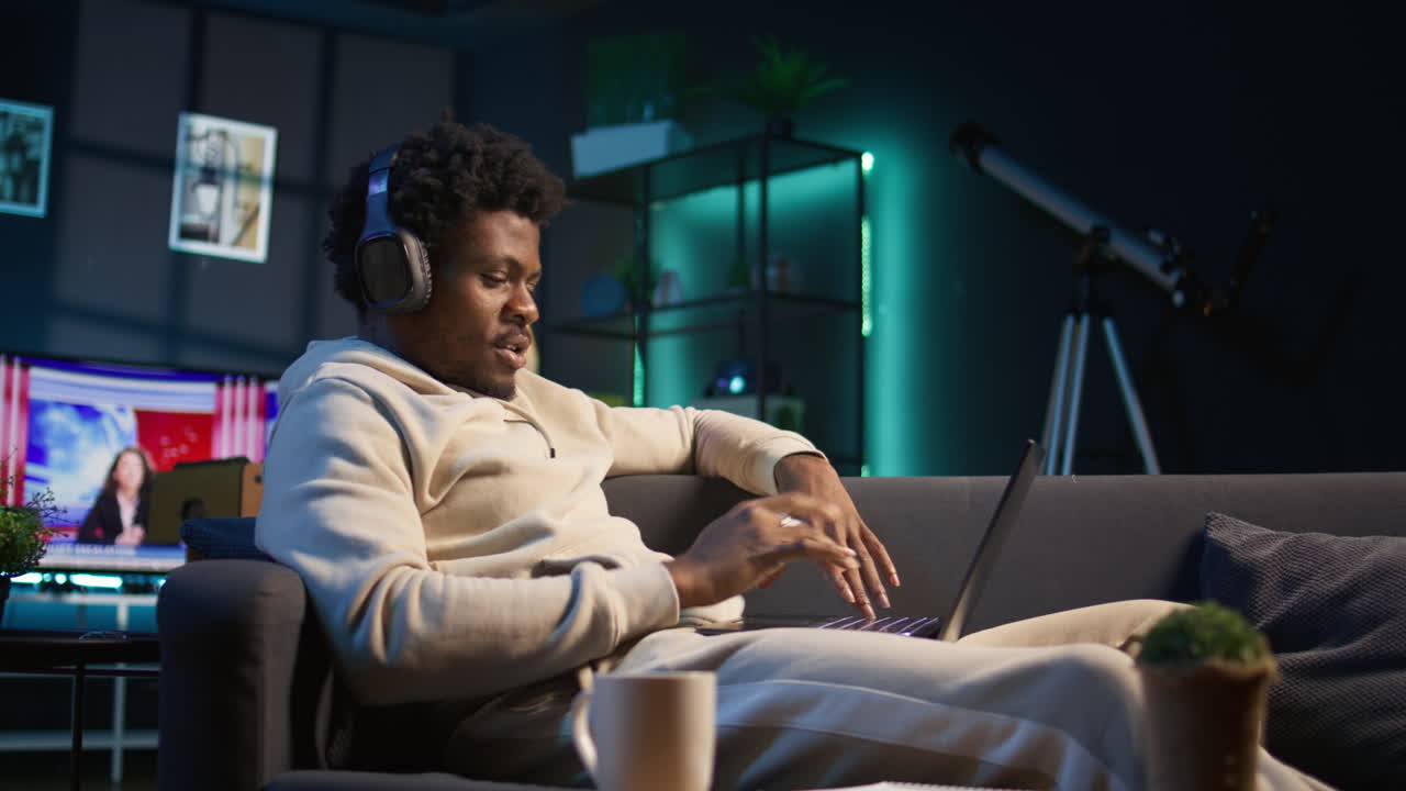 Freelancer listening to a podcast and being focused on online work, putting on headphones while seated on the couch in his living room. Productivity in a casual and flexible home. Camera B.