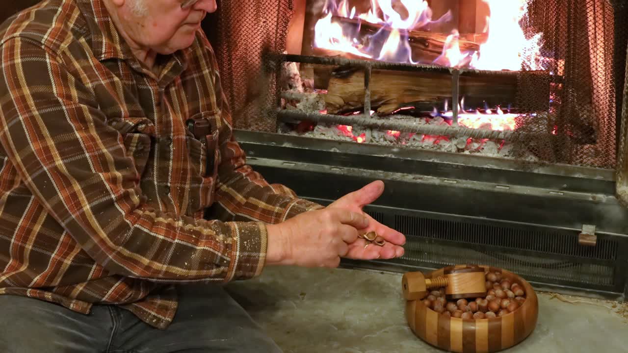 el anciano está usando una galleta de nueces hecha a mano para abrir avellanas mientras se sienta en la chimenea