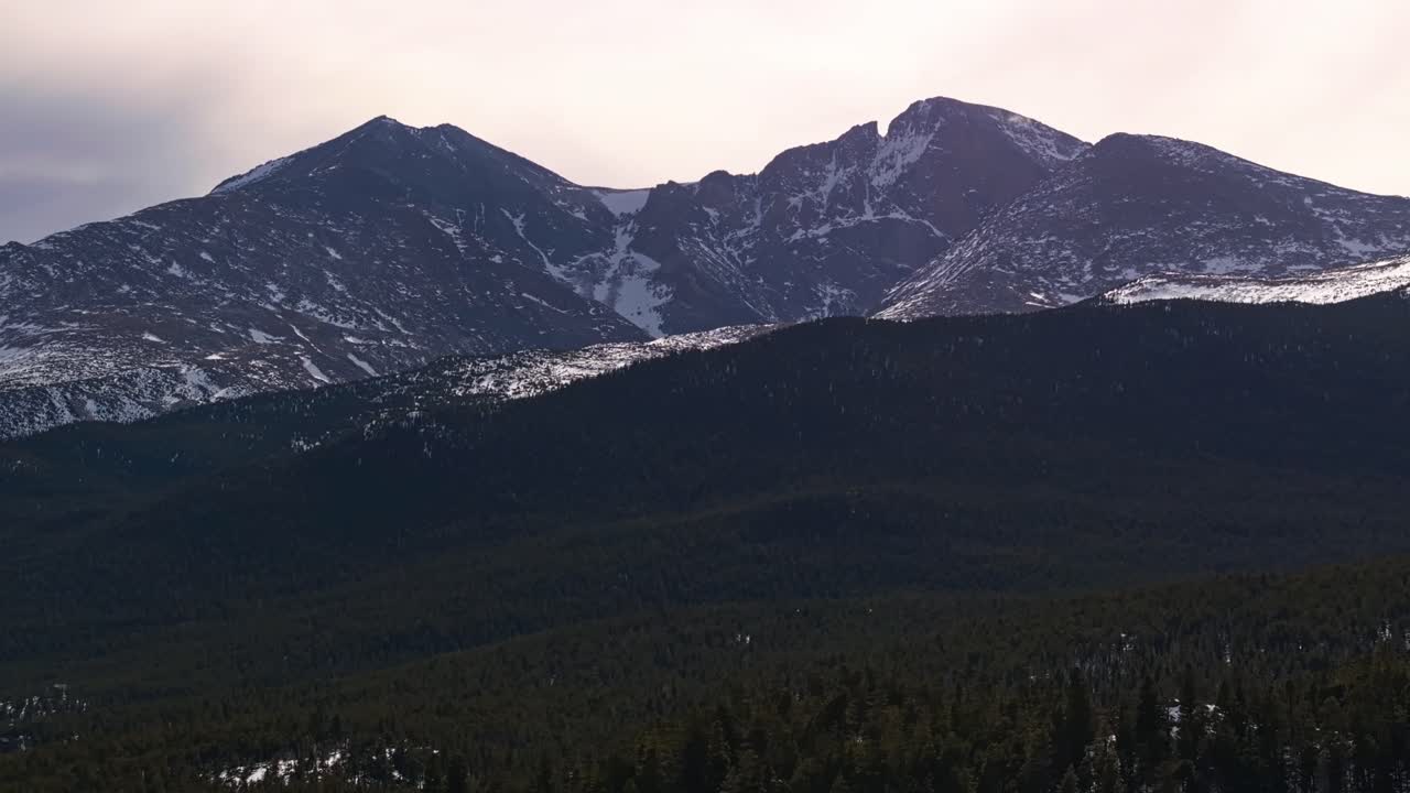 Trees and snow patches dot a sloped clearing beneath rocky cliffs and overcast sky, tracking left aerial, Allenspark Colorado USA