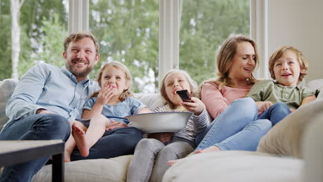 Familie-Sitzt-Zu-Hause-Auf-Dem-Sofa,-Lacht-Und-Schaut-Zusammen-Mit-Popcorn-Fern