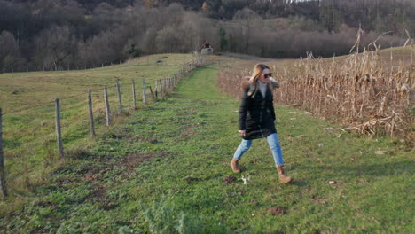 Young-white-woman-model-walking-in-old-small-village-with-amazing-agricultural-field-and-stunning-nature-with-green-grass-and-forest-in-background-with-small-old-traditional-house