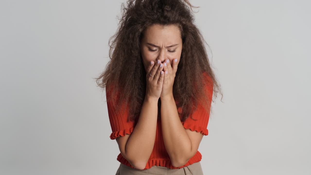 una mujer caucásica de cabello rizado tosendo frente a la cámara.