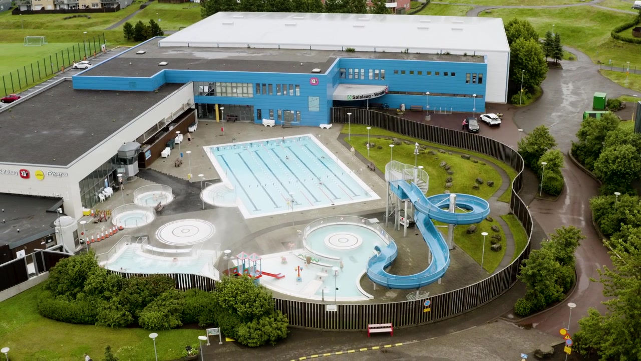 High angle aerial view of a large public swimming pool complex in Iceland, with people swimming in outdoor geothermal pools, hot tubs, and using a large blue water slide.