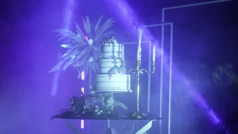 Dramatisch-Beleuchtete-Hochzeitstorte-Mit-Floralen-Akzenten-Und-Kerzen-In-Einer-Violett-blauen-Nebligen-Atmosphäre