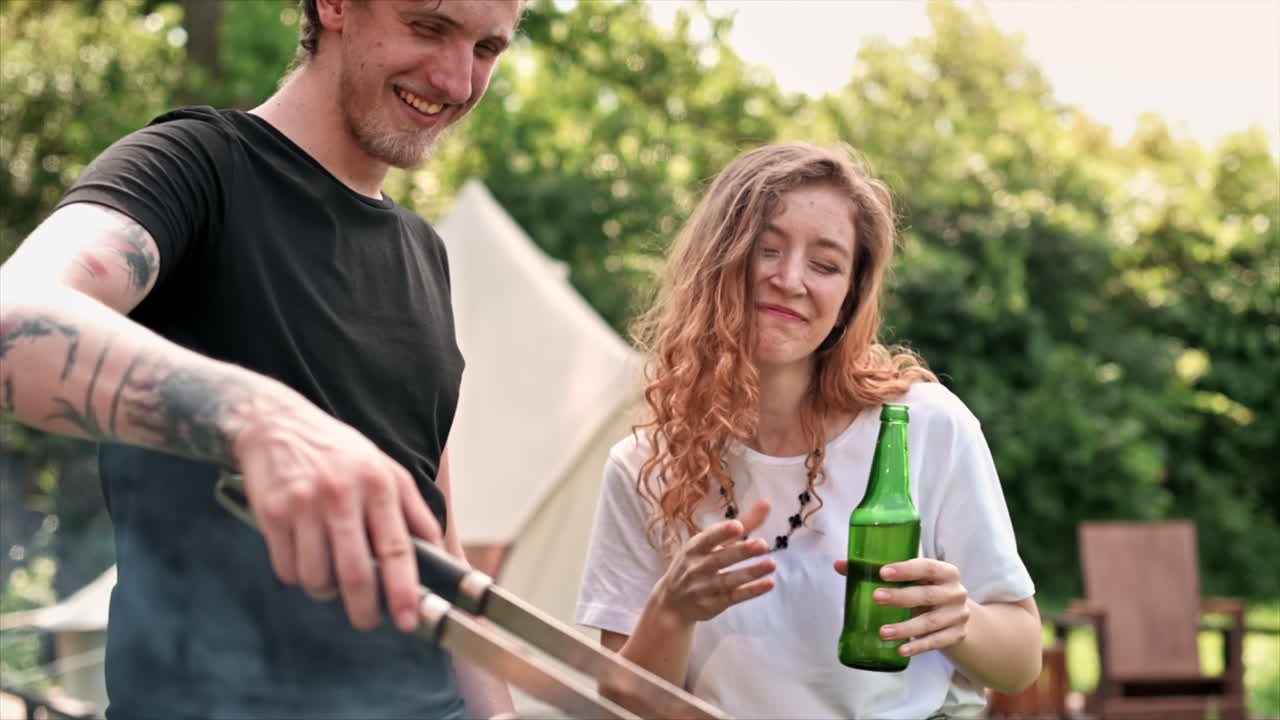Young couple frying meat on the grill and drinking beer, laughing. Greenery around. Glamping. Slow motion