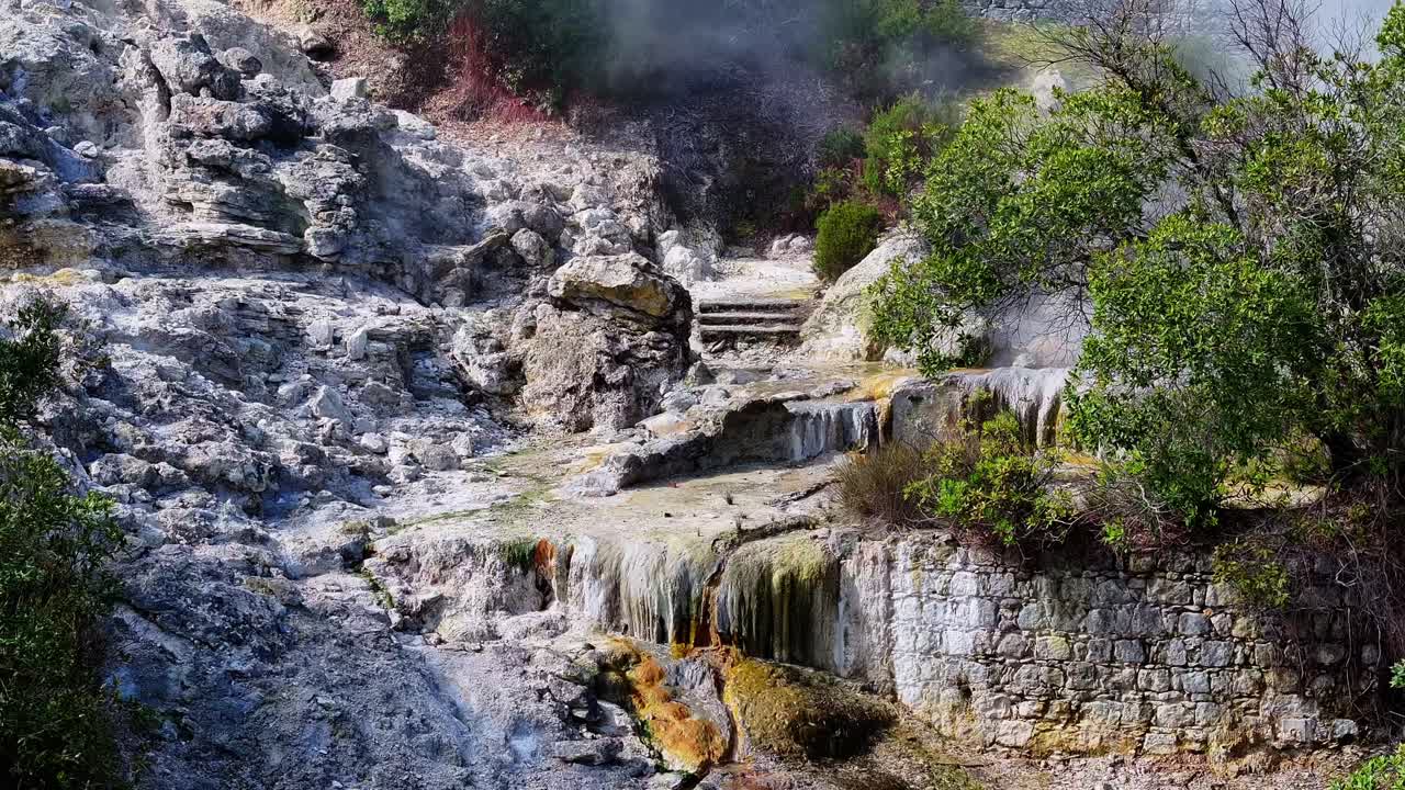 มุมมองทางอากาศของปล่องฟูมารอล Furnas ที่ซึ่งไอน้ำพวยพุ่งขึ้นจากช่องระบายความร้อนใต้พิภพ ล้อมรอบด้วยหินสีเข้มและพื้นผิวภูเขาไฟที่ตัดกันในหมู่เกาะอะซอเรส ประเทศโปรตุเกส