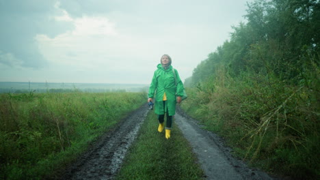 mujer con impermeable verde llevando bolsa de color menta y paraguas, con botas de lluvia amarillas, caminando por un camino fangoso rodeado de vegetación y árboles bajo un cielo brumoso