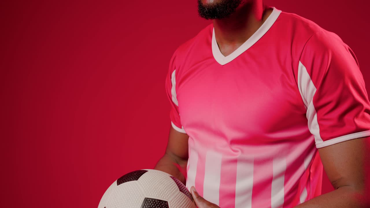 Hombre africano con camiseta rosa ajustando un balón de fútbol en estudio, la cámara se aleja para un retrato, espacio para texto