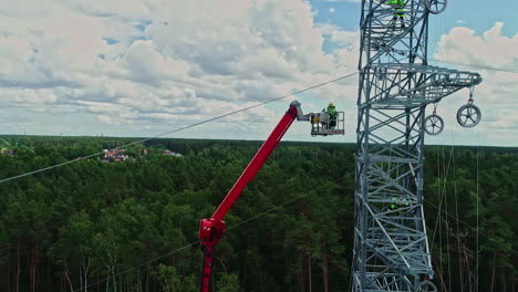 static aerial drone shot of one among the three technicians is coming down using lifting crane, other two are staying up while working on high voltage electricity tower in their uniform