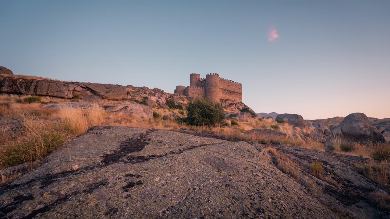 Timelapse of the medieval Manqueospese Castle in Ávila, Spain, bathed in golden sunset light. Rocky foreground and warm tones create a cinematic historic landscape