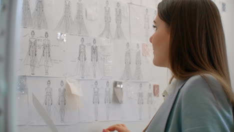 young fashion designer standing with back to camera looking at drawings sketches hanging on wall. woman is thinking about new trendy clothing collection