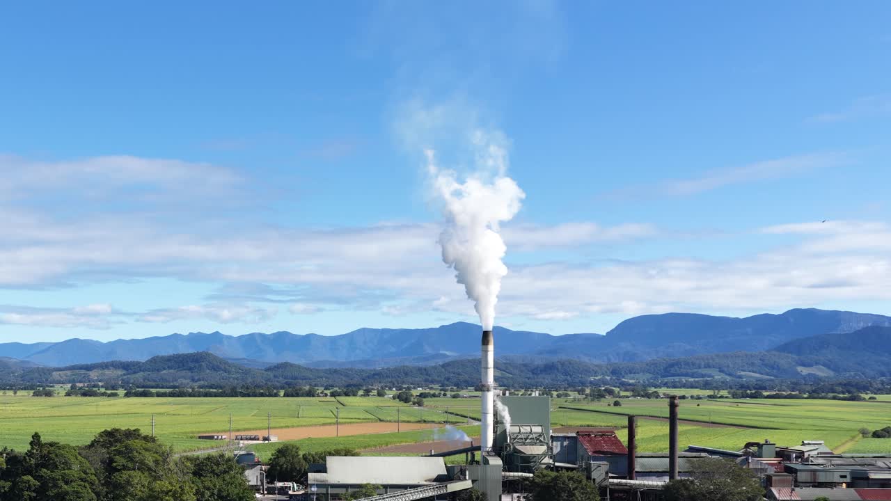 Aerial view of a sugar mill with steam rising, surrounded by lush landscapes and mountains under clear skies