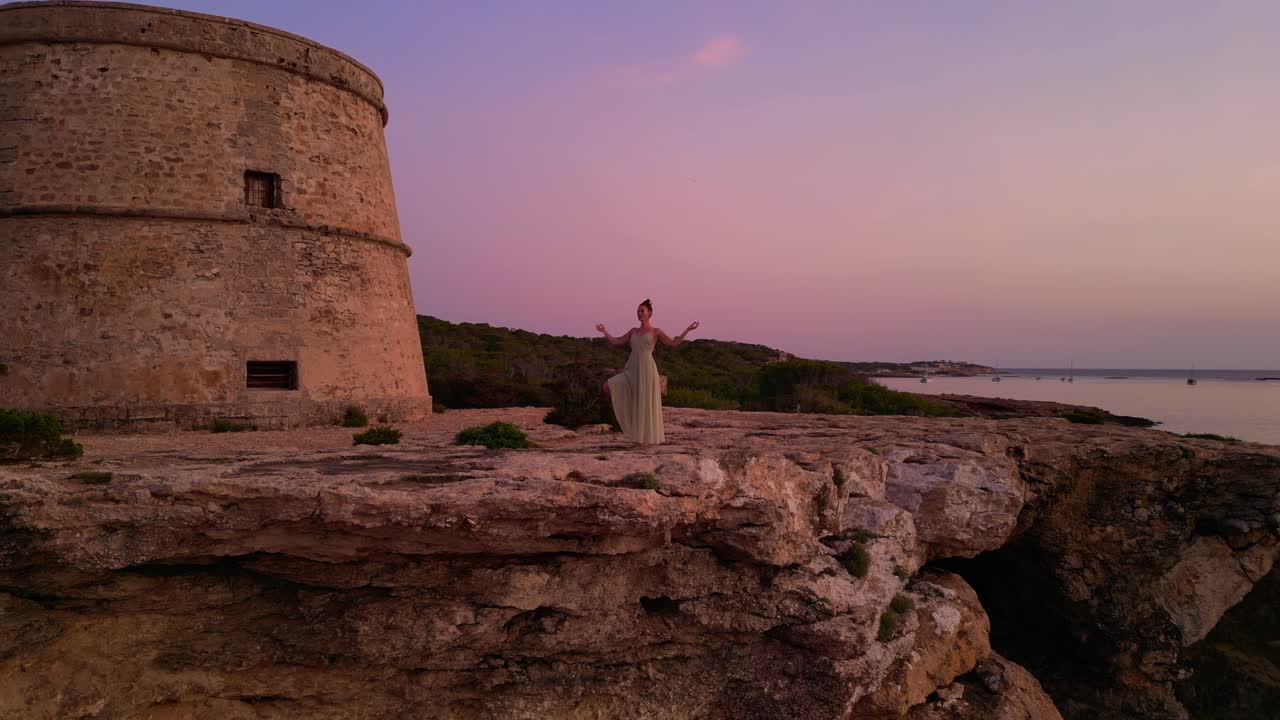 Donna che esegue posizioni yoga al tramonto vicino alla Torre e all'isola di Es Vedrà a Ibiza, Spagna. Bella ripresa aerea con drone in discesa.