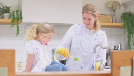 Madre-Usando-Guantes-De-Goma-En-Casa-En-La-Cocina-Con-Su-Hija-Pequeña-Divirtiéndose-Mientras-La-Niña-Pone-Los-Pies-En-El-Fregadero-Mientras-Ellos-Lavan-En-El-Fregadero,-Filmada-En-Cámara-Lenta