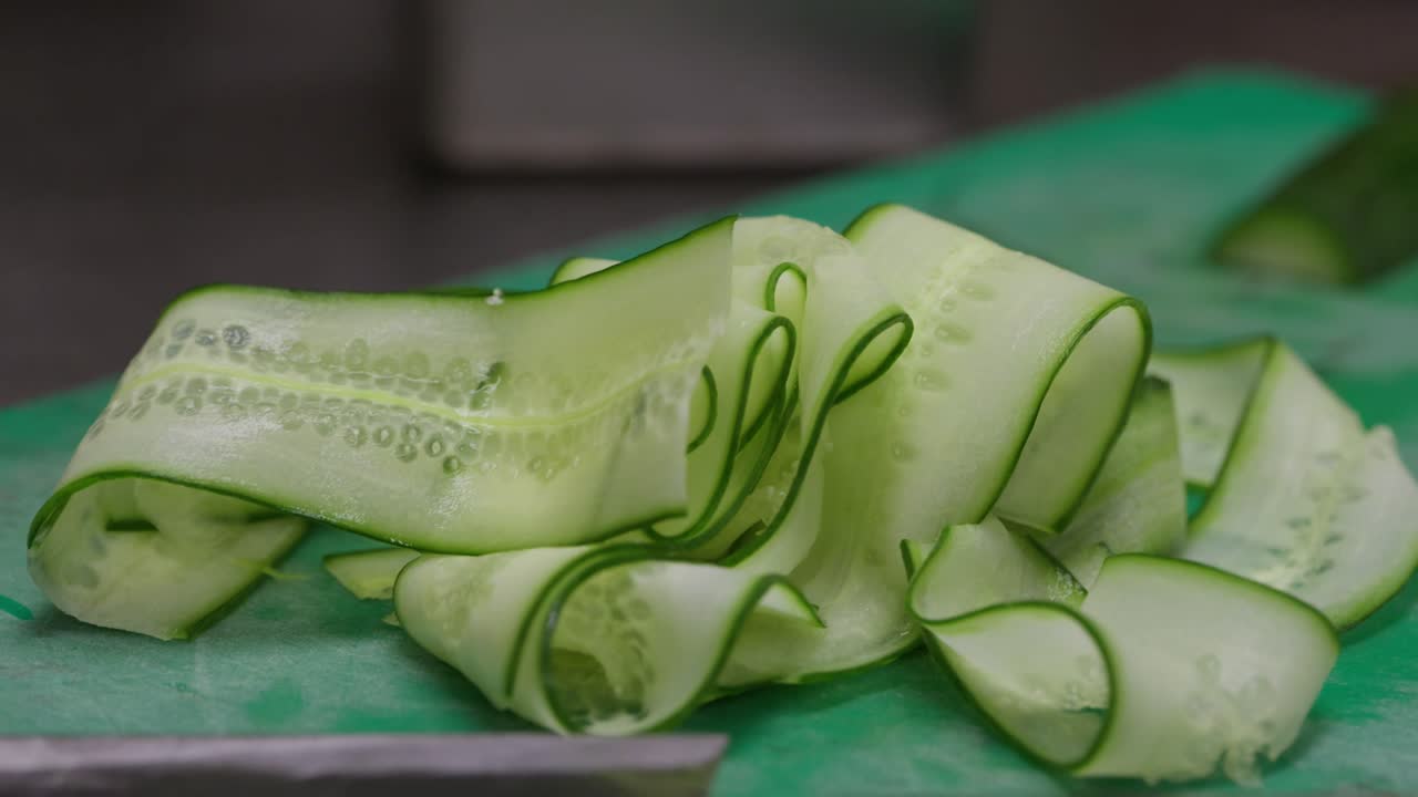 Zoom in close up of a pile of cucumber ribbons and a wooden handled knife on a green chopping board