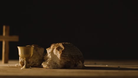 Religious-Concept-Shot-With-Chalice-Broken-Bread-Cross-And-Wine-On-Wooden-Table-Against-Black-Background-1