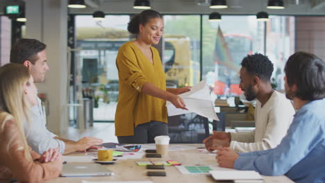 businesswoman giving presentation to colleagues sitting around table in modern open plan office