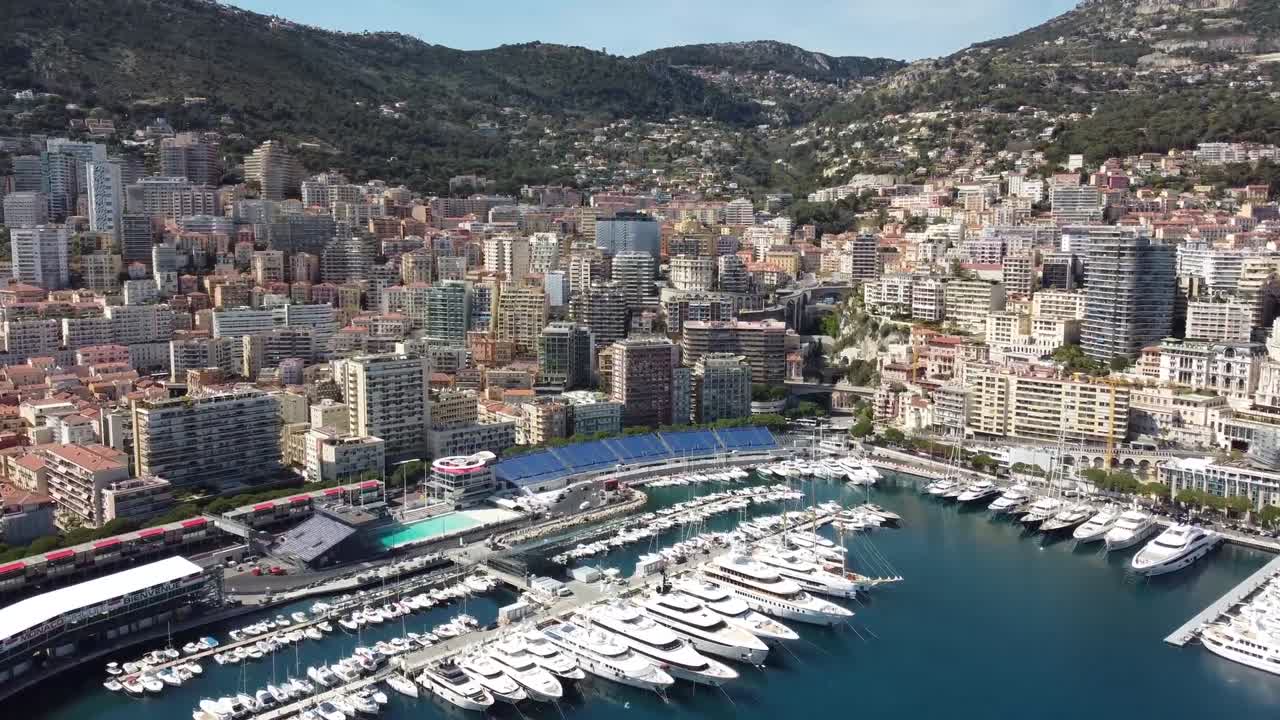 Aerial drone footage of Port Hercules in Monaco showing luxury yachts docked along the marina with hillside buildings rising above capturing Mediterranean elegance, architecture, and wealth