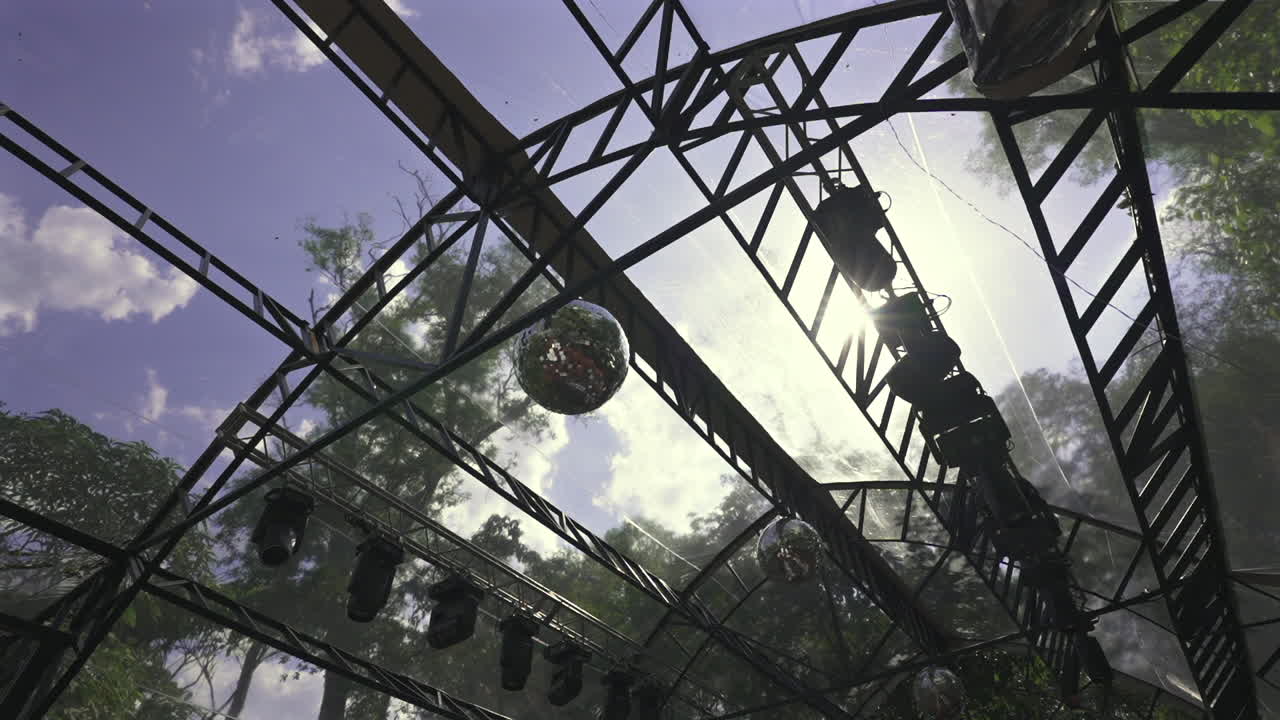SA slow pan reveals a transparent gazebo roof adorned with disco balls and lights, set in a jungle wedding venue where metal trusses meet tropical trees and filtered sunlight, ready for events